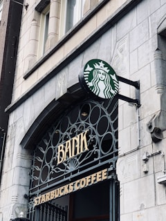 A Starbucks coffee shop sign is displayed on an ornate building facade that features the word 'BANK' just above the entrance. The building is constructed with stone and features architectural details, including carved elements and ironwork.