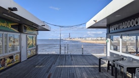 A wooden boardwalk leads to a scenic view of a sandy beach with a shoreline and distant buildings. The sky is clear and blue. On either side of the walkway, there are small food stalls with brightly colored signs, one offering Caribbean cuisine and another featuring seafood.
