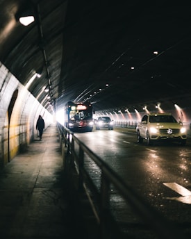 A dimly lit tunnel features vehicles like a bus and cars driving through. On the left side, a pedestrian walks along the sidewalk, with light emanating from the tunnel's end, creating a sense of depth and motion.