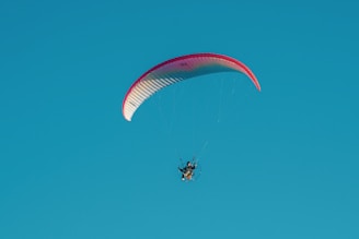 A paraglider with a red and white canopy is soaring against a clear blue sky. The pilot is visible, controlling the glider through a harness and motorized propeller.
