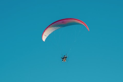 A paraglider with a red and white canopy is soaring against a clear blue sky. The pilot is visible, controlling the glider through a harness and motorized propeller.