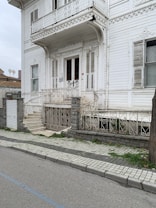A detailed view of an old, ornate white wooden house with intricate carvings and decorative elements. The house has a small, raised porch and a staircase leading to the entrance, with metal railings. The structure features several windows with shutters, some partially open, and there are signs of weathering on the wood. The surrounding area includes a stone fence and a tiled sidewalk with patches of grass.