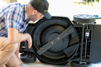 A person in a plaid shirt and cap is inspecting the underside of a lawnmower. The lawnmower is flipped over, revealing its large circular blade in the center. The lawnmower has large plastic wheels and a sturdy frame.