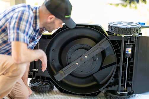 A person in a plaid shirt and cap is inspecting the underside of a lawnmower. The lawnmower is flipped over, revealing its large circular blade in the center. The lawnmower has large plastic wheels and a sturdy frame.