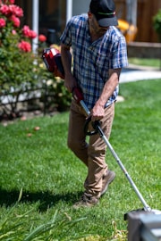 A person is engaged in yard work, using a grass trimmer on a well-maintained lawn. They are wearing a plaid shirt, beige pants, and a black cap. To the side, there are blooming red flowers and a glimpse of a house with windows and a wooden fence.