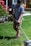 A person is engaged in yard work, using a grass trimmer on a well-maintained lawn. They are wearing a plaid shirt, beige pants, and a black cap. To the side, there are blooming red flowers and a glimpse of a house with windows and a wooden fence.
