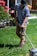 A person is engaged in yard work, using a grass trimmer on a well-maintained lawn. They are wearing a plaid shirt, beige pants, and a black cap. To the side, there are blooming red flowers and a glimpse of a house with windows and a wooden fence.