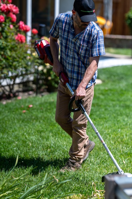A person is engaged in yard work, using a grass trimmer on a well-maintained lawn. They are wearing a plaid shirt, beige pants, and a black cap. To the side, there are blooming red flowers and a glimpse of a house with windows and a wooden fence.
