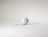 A single white golf ball with a textured dimple pattern rests on a plain, light-colored surface, casting a subtle shadow.