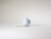 A single white golf ball with a textured dimple pattern rests on a plain, light-colored surface, casting a subtle shadow.