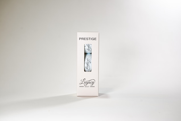A package labeled 'Prestige' containing a single golf ball with the word 'Legacy' written on it. The packaging is white with a transparent section displaying the ball. The overall design is minimalist and sleek.