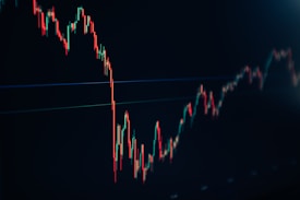 A financial chart displaying a downward trend with multiple sharp peaks and valleys. The chart features predominantly red and green candlesticks against a dark background, indicating fluctuations in market values.