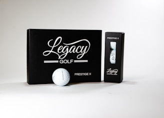 A neatly arranged display featuring a box and a sleeve with 'Legacy Golf' branding. The larger black box is labeled 'Prestige X' and is complemented by a golf ball in front of it. A smaller sleeve containing additional golf balls sits to the right. The design conveys sophistication and quality.