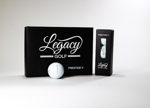 A neatly arranged display featuring a box and a sleeve with 'Legacy Golf' branding. The larger black box is labeled 'Prestige X' and is complemented by a golf ball in front of it. A smaller sleeve containing additional golf balls sits to the right. The design conveys sophistication and quality.