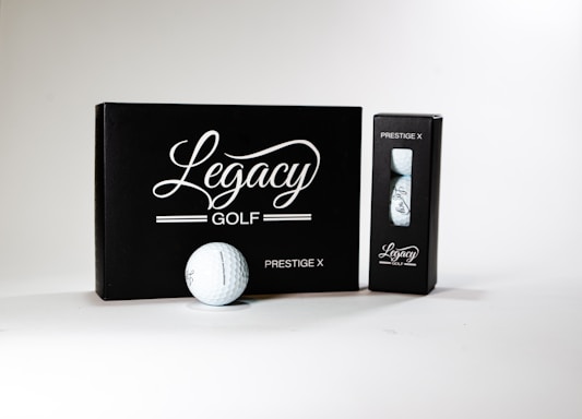 A neatly arranged display featuring a box and a sleeve with 'Legacy Golf' branding. The larger black box is labeled 'Prestige X' and is complemented by a golf ball in front of it. A smaller sleeve containing additional golf balls sits to the right. The design conveys sophistication and quality.