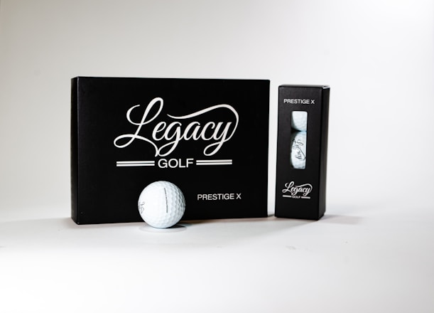A neatly arranged display featuring a box and a sleeve with 'Legacy Golf' branding. The larger black box is labeled 'Prestige X' and is complemented by a golf ball in front of it. A smaller sleeve containing additional golf balls sits to the right. The design conveys sophistication and quality.