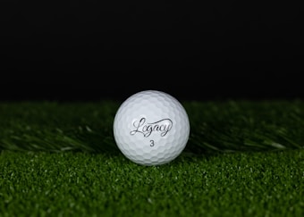 A golf ball with the word 'Legacy' and the number '3' printed on it rests on a patch of green grass. The background is dark, providing contrast to the white of the golf ball.