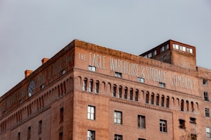 A large, historic brick building with 'The Eagle Warehouse & Storage Company' written prominently in white letters across the top. The structure features arched windows and decorative brickwork, adding to its architectural interest. A smaller section on top looks to be more modern, with larger windows overlooking the view.