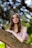 A young woman with long brown hair poses gracefully against a tree branch. She is wearing a delicate, pink, off-the-shoulder gown. Her makeup is elaborate with dark eyelashes and small jewels around her eyes. The background is a blur of bright, natural greenery, creating a dreamy bokeh effect.