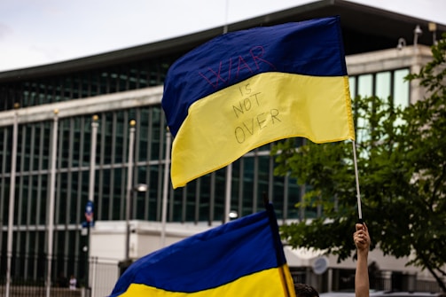 A blue and yellow flag with the words 'WAR IS NOT OVER' written on it, held up by a person in front of a modern building with large windows. There is another similar flag in the foreground and some greenery is visible on the right.