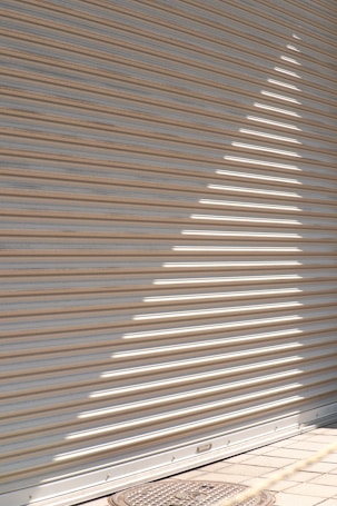 A corrugated metal shutter with horizontal lines is partially illuminated by sunlight, creating a bright triangular pattern. The light forms a sharp contrast against the shaded areas, and a round metal manhole cover is visible at the bottom on a tiled pavement.