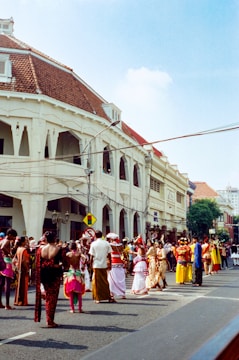 A lively street scene with a group of people dressed in traditional, colorful costumes participating in what appears to be a cultural parade or festival. The architecture of the surrounding buildings, featuring white colonial-style facades and red-tiled roofs, adds to the vibrant atmosphere.