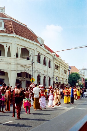 A lively street scene with a group of people dressed in traditional, colorful costumes participating in what appears to be a cultural parade or festival. The architecture of the surrounding buildings, featuring white colonial-style facades and red-tiled roofs, adds to the vibrant atmosphere.