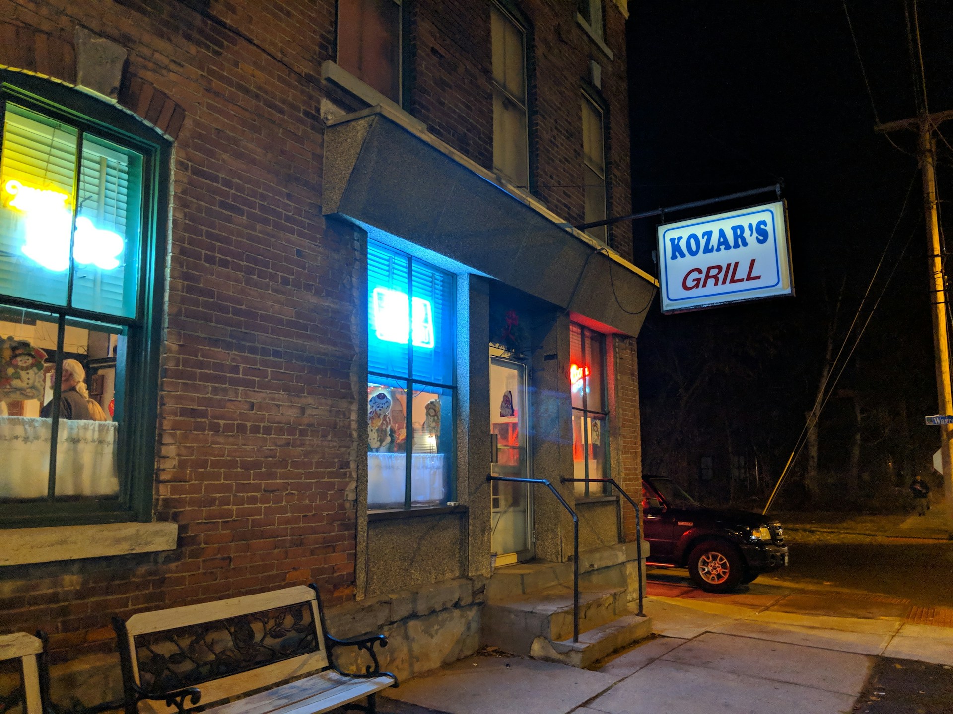 A nighttime scene featuring the exterior of a brick building with a sign for 'Kozar's Grill' illuminated in white and red neon lights. Bright blue and yellow neon signs also light up the windows, and a small black SUV is parked on the street nearby. A bench with decorative metal armrests is positioned against the building.