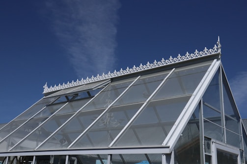 A greenhouse structure with a pitched glass roof decorated with ornate white trim against a clear blue sky. The roof consists of multiple glass panels supported by white metal framing, and an intricate white decorative ridge adorns its top.