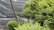 A lush, green garden scene featuring carefully manicured shrubs and small trees. The plants display a dense and vibrant foliage, with the branches artfully pruned, suggesting attentive gardening practice. A large net or mesh structure serves as a backdrop, possibly providing protection or shade to the garden.