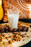 A glass of milk is placed on a wooden board surrounded by an assortment of nuts and dates. In the background, there is a colorful pillow with intricate patterns in orange and white.