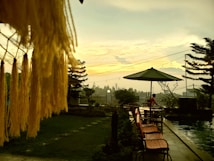 A tranquil outdoor scene featuring a swimming pool, a line of wooden lounge chairs, and a large green umbrella. In the foreground, yellow tassels hang down, creating a cozy atmosphere. The background is adorned with a string of lights, silhouetted pine trees, and a person standing near the pool, gazing into the distance during a sunset with a soft, cloudy sky.