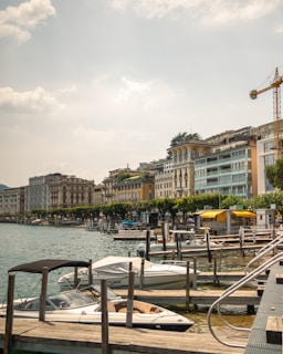 A picturesque lakeside view featuring a row of elegant, multi-story buildings with balconies, set against a backdrop of gently rolling hills. A series of moored boats and yachts line the wooden docks in the foreground, while a yellow canopy stands out among the lush, green trees. A construction crane is visible to the right, indicating ongoing development.