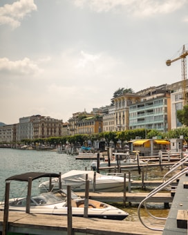 A picturesque lakeside view featuring a row of elegant, multi-story buildings with balconies, set against a backdrop of gently rolling hills. A series of moored boats and yachts line the wooden docks in the foreground, while a yellow canopy stands out among the lush, green trees. A construction crane is visible to the right, indicating ongoing development.
