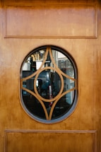 A wooden door with an oval-shaped glass insert featuring a decorative geometric wooden design in the center. Through the glass, various objects and furniture, possibly in a room or shop, are visible in the background.