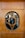 A wooden door with an oval-shaped glass insert featuring a decorative geometric wooden design in the center. Through the glass, various objects and furniture, possibly in a room or shop, are visible in the background.