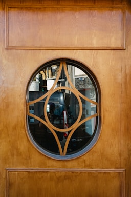 A wooden door with an oval-shaped glass insert featuring a decorative geometric wooden design in the center. Through the glass, various objects and furniture, possibly in a room or shop, are visible in the background.