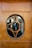 A wooden door with an oval-shaped glass insert featuring a decorative geometric wooden design in the center. Through the glass, various objects and furniture, possibly in a room or shop, are visible in the background.