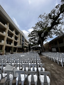 A large outdoor venue features rows of white plastic chairs aligned in a semi-circle facing a stage. The setting is between two large buildings, with a massive tree to the right providing partial shade. The sky is overcast, contributing to a subdued ambiance.
