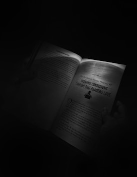 An open book held by someone's hands is illuminated by a focused light source in an otherwise dark environment. One page features a heading 'Creating Consistent Content That Readers Love' and some text below it.