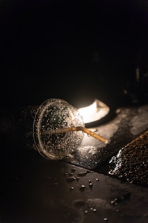 A disposable plastic cup with a straw lies on a dark, wet surface, illuminated by a single light source in the background. Water droplets are visible on the cup and surrounding surface, creating a reflective effect.