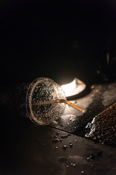 A disposable plastic cup with a straw lies on a dark, wet surface, illuminated by a single light source in the background. Water droplets are visible on the cup and surrounding surface, creating a reflective effect.