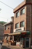 A small convenience store with a brick facade is situated in an urban area. The store has a colorful awning with red, green, and orange stripes. Above the entrance, there is a sign with Korean writing. Various items such as boxes and containers are placed outside the store. A vending machine is also to the right of the entrance. The building is three stories high with multiple windows, and there is another similar brick building adjacent to it.