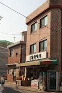 A small convenience store with a brick facade is situated in an urban area. The store has a colorful awning with red, green, and orange stripes. Above the entrance, there is a sign with Korean writing. Various items such as boxes and containers are placed outside the store. A vending machine is also to the right of the entrance. The building is three stories high with multiple windows, and there is another similar brick building adjacent to it.