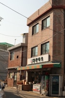 A small convenience store with a brick facade is situated in an urban area. The store has a colorful awning with red, green, and orange stripes. Above the entrance, there is a sign with Korean writing. Various items such as boxes and containers are placed outside the store. A vending machine is also to the right of the entrance. The building is three stories high with multiple windows, and there is another similar brick building adjacent to it.