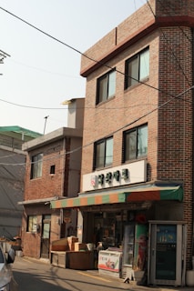 A small convenience store with a brick facade is situated in an urban area. The store has a colorful awning with red, green, and orange stripes. Above the entrance, there is a sign with Korean writing. Various items such as boxes and containers are placed outside the store. A vending machine is also to the right of the entrance. The building is three stories high with multiple windows, and there is another similar brick building adjacent to it.