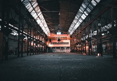 An abandoned industrial warehouse with a large, open floor space. The structure features rusted metal beams and a ceiling with long skylights that let in natural light. The far end of the warehouse has a windowed room elevated above the floor, possibly an office space, with a staircase leading to it. Graffiti adorns the walls, adding a touch of color and urban decay.