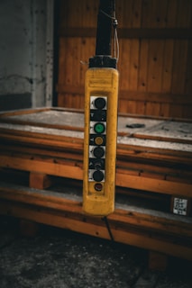 A close-up view of a hanging industrial control switch with multiple buttons, encased in a yellow protective cover. The switch is situated in an industrial setting, possibly in a workshop, with a wooden pallet and a partially visible door in the background.