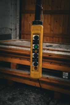 A close-up view of a hanging industrial control switch with multiple buttons, encased in a yellow protective cover. The switch is situated in an industrial setting, possibly in a workshop, with a wooden pallet and a partially visible door in the background.