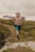 A young girl is energetically jumping across a puddle on a trail surrounded by grassy terrain. She is wearing a pink hoodie, turquoise leggings, and dark shoes. Her determined expression and the motion of her jump suggest excitement and playfulness.